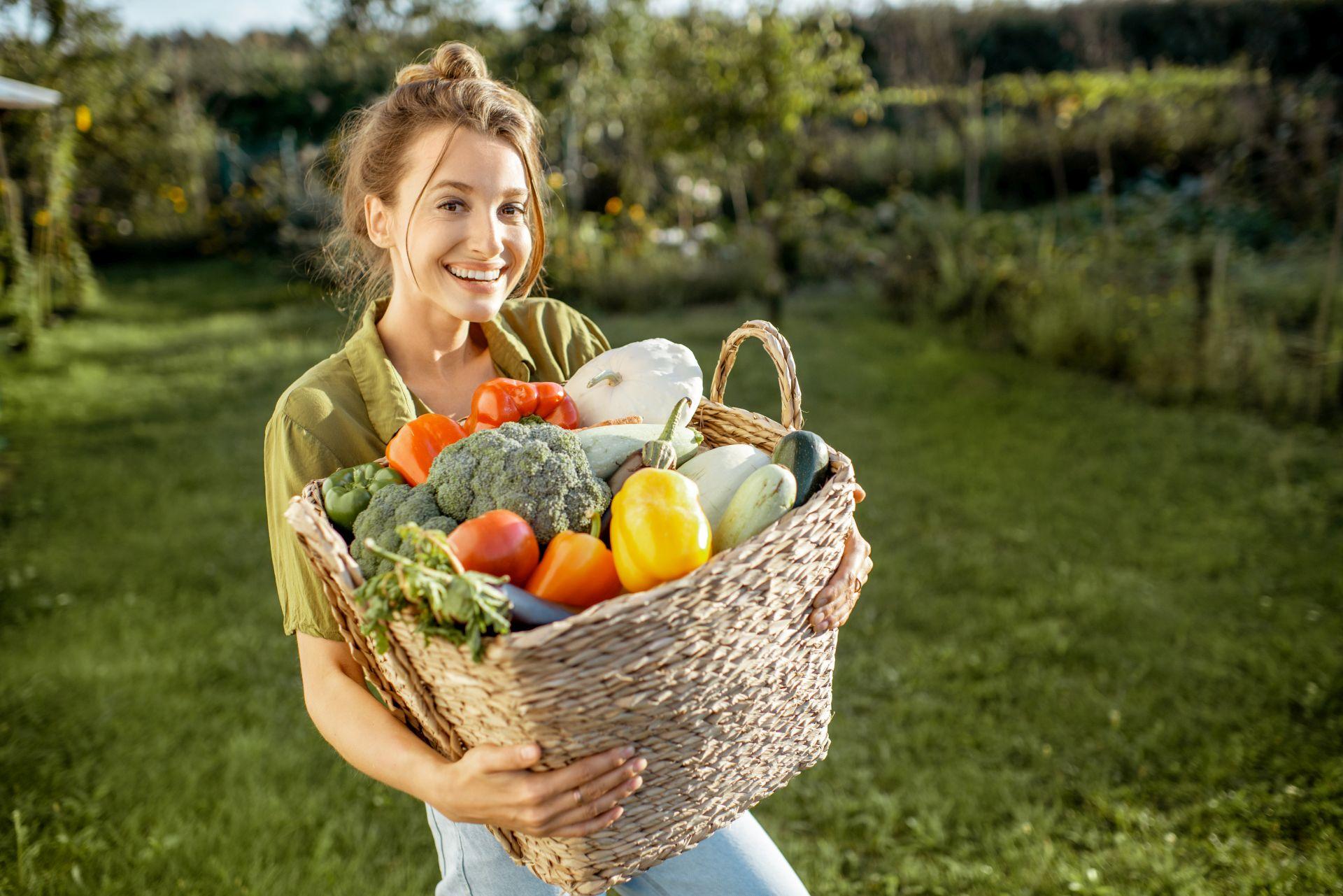 Eine lächelnde Frau in grüner Kleidung hält einen Korb mit frischem Bio-Gemüse wie Paprika, Brokkoli und Zucchini. Im Hintergrund ist ein üppiger Garten zu sehen, der den Fokus auf natürliche und nachhaltige Ernährung legt.