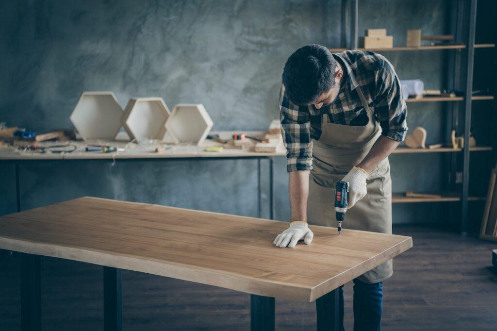 Handwerker bei der Arbeit an einem Holzesstisch. Mit Bohrer und Schutzhandschuhen veredelt er die Tischplatte in einer modernen Werkstatt voller Holzprojekte.